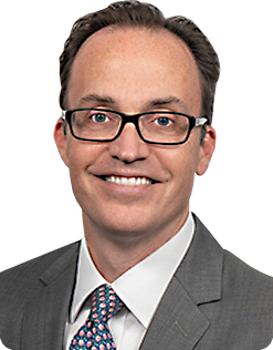 Professional headshot of a smiling man in a gray suit, white shirt, and patterned tie, wearing glasses.